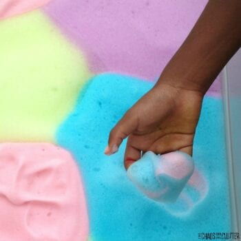 a black child's hand is reaching up from a bin of soap foam holding blue and pink soap foam. The bin contains areas of pink, blue, purple, and yellow soap foam.