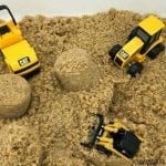 a white bin filled with kinetic sand also contains 3 small construction vehicle toys that are yellow and black in colour.