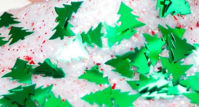 an almost opaque white slime with red round confetti and bright green tree shaped confetti is seen held in a hand with only the thumb visible