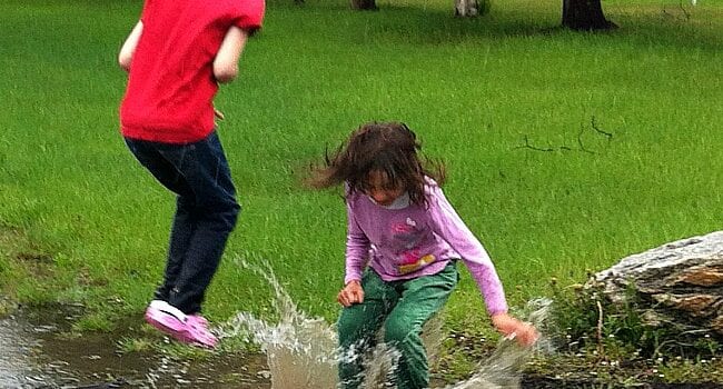 There are trees in the background and bright green grass. Two young girls, one wearing a red t-shirt and black pants with pink shoes, the other wearing a pink long sleeved shirt and green pants are splashing in a puddle. The water splash comes up higher than the girl in the green pants' knees. The other girl is jumping in the air.