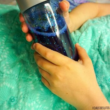 a child's hands hold a blue calm down bottle. The child's hands are all that is visible as the child is snuggled underneath a green blanket.