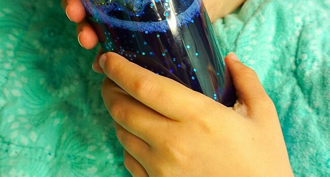a child's hands hold a blue calm down bottle. The child's hands are all that is visible as the child is snuggled underneath a green blanket.