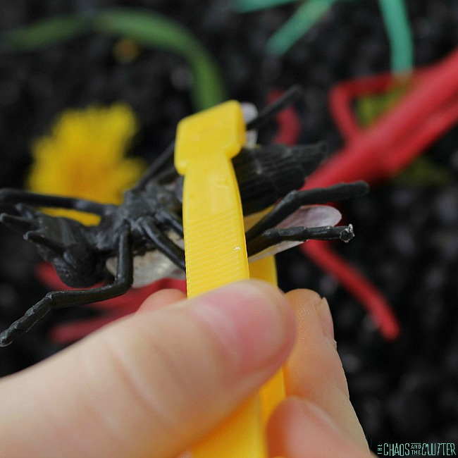 a child hand pinching yellow tweezers that are holding a black plastic bug
