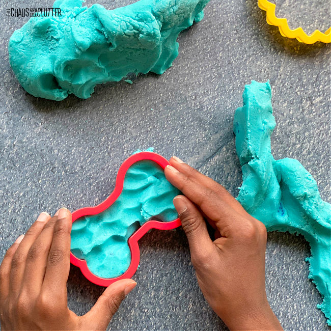 child's hands press a dog bone shaped cookie cutter into blue playdough