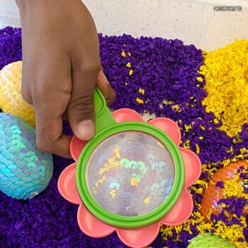 child's hand holding a magnifying glass over a sequined egg
